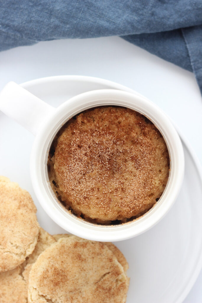 top down image of a white mug filled with baked cinnamon cake on a white plate with snickerdoodle cookies on the side. 