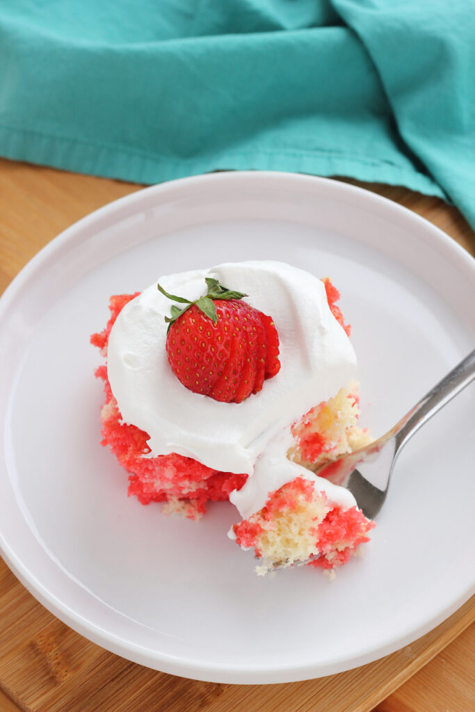 white round plate with a slight ridge on the edges with a single slice of red and white cake that is topped with cool whip and a fresh strawberry that is sliced. One bite is sitting on a fork. 