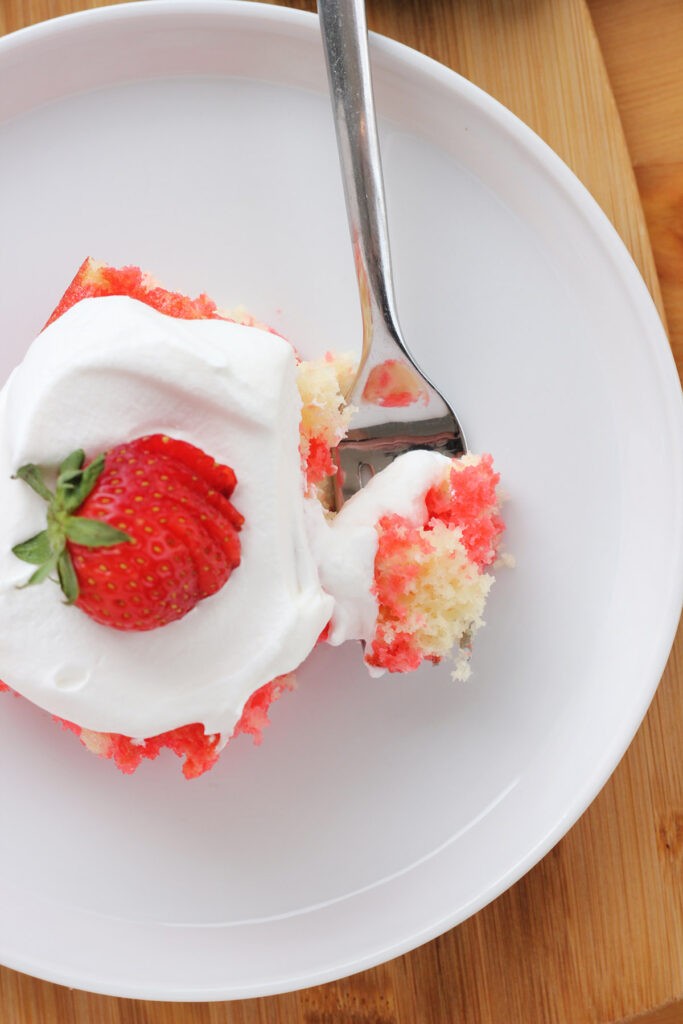 top down image of a white round plate that is sitting on a wooden cutting board. On top of the plate is a single slice of poke cake with a bite sitting on a fork and a sliced berry on the top