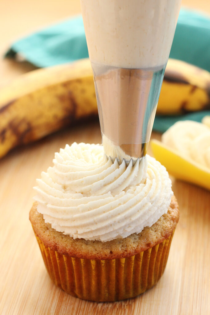 white frosting being pipped onto a cupcake in a yellow baking liner. The cupcake is sitting on a wooden tabletop with overripe bananas sitting in the background with a teal napkin