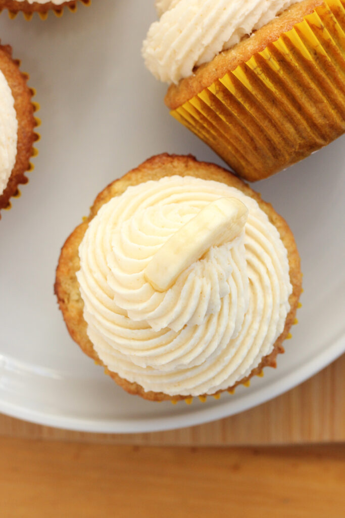 top down image of a cupcake that is sitting on a white plate with pipped white frosting and a fresh banana slice. Other cupcakes are scattered around the edge of the plate