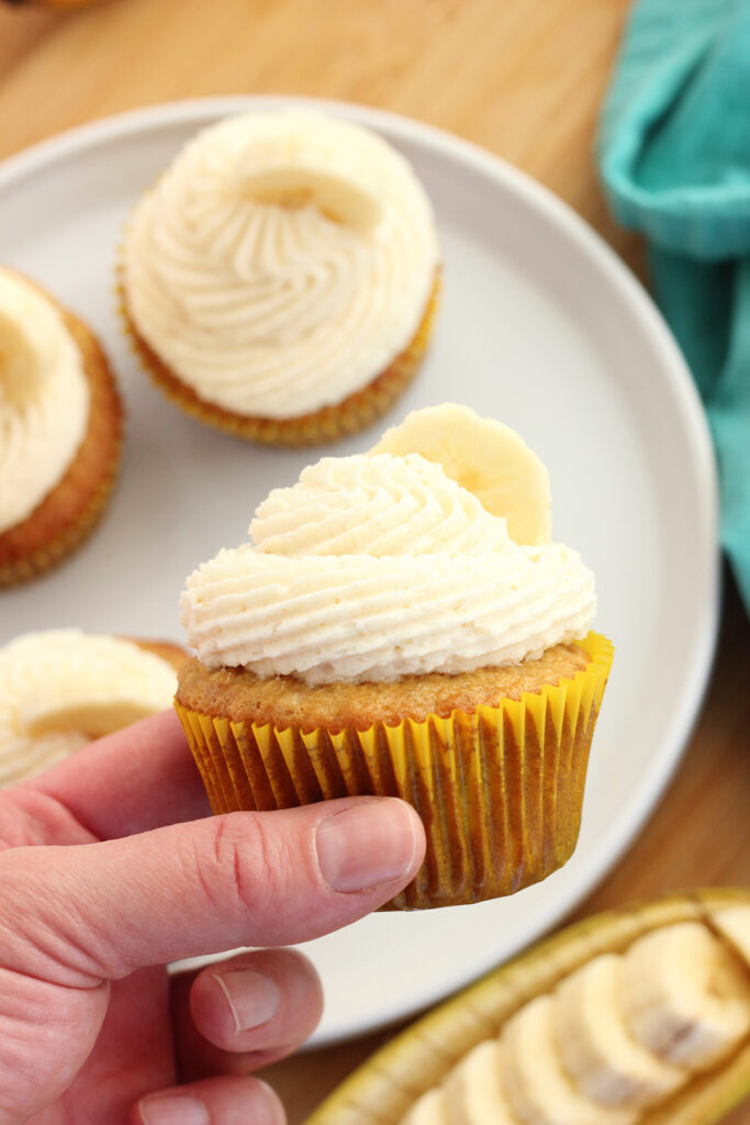 hand holding a cupcake in a yellow wrapper with white pipped frosting and a banana slice on top. Hovering over a white plate with more cupcakes and a wooden table with teal napkin off to the edge of the frame