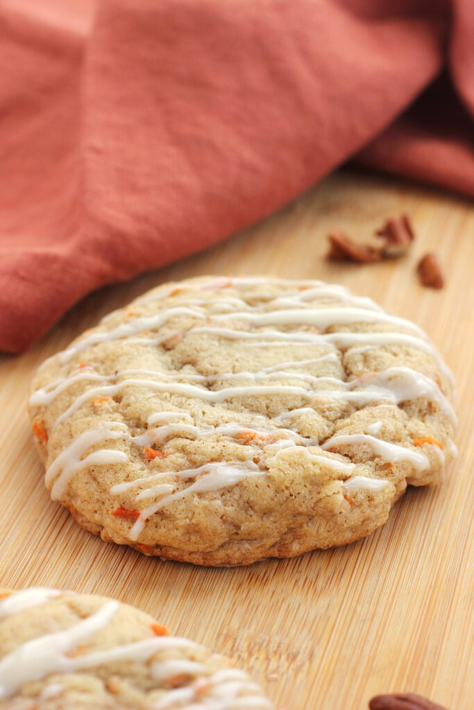 close up image of a carrot cookie with a drizzle of cream cheese glaze sitting on a wooden cutting board with a dark orange napkin in the background