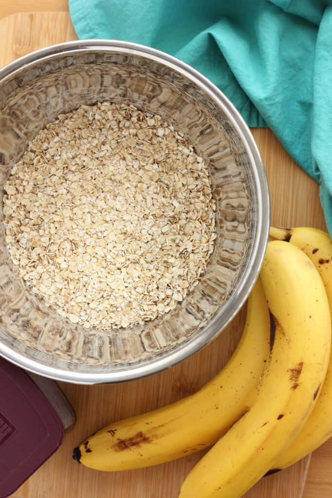 silver mixing bowl filled with quick oats sitting on a wooden cutting board with a side of bananas and a teal napkin