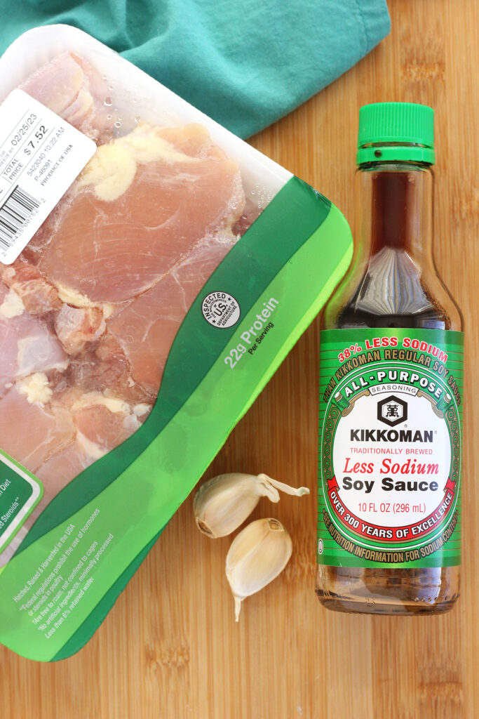wooden cutting board topped with a bottle of low sodium soy sauce, container of chicken thighs, and two garlic cloves