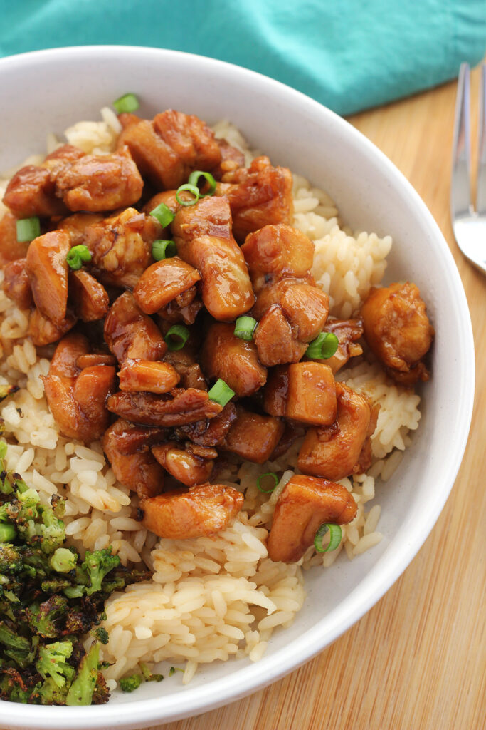 Close up image of chicken with teriyaki sauce sitting on a bed of rice with green onion sprinkled on top. All of this is in a white bowl that is on a wooden table with a teal napkin