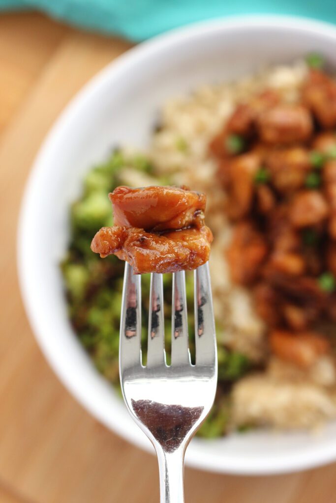 close up image of a bite of teriyaki chicken on a fork hovering over a bowl of chicken, rice and broccoli