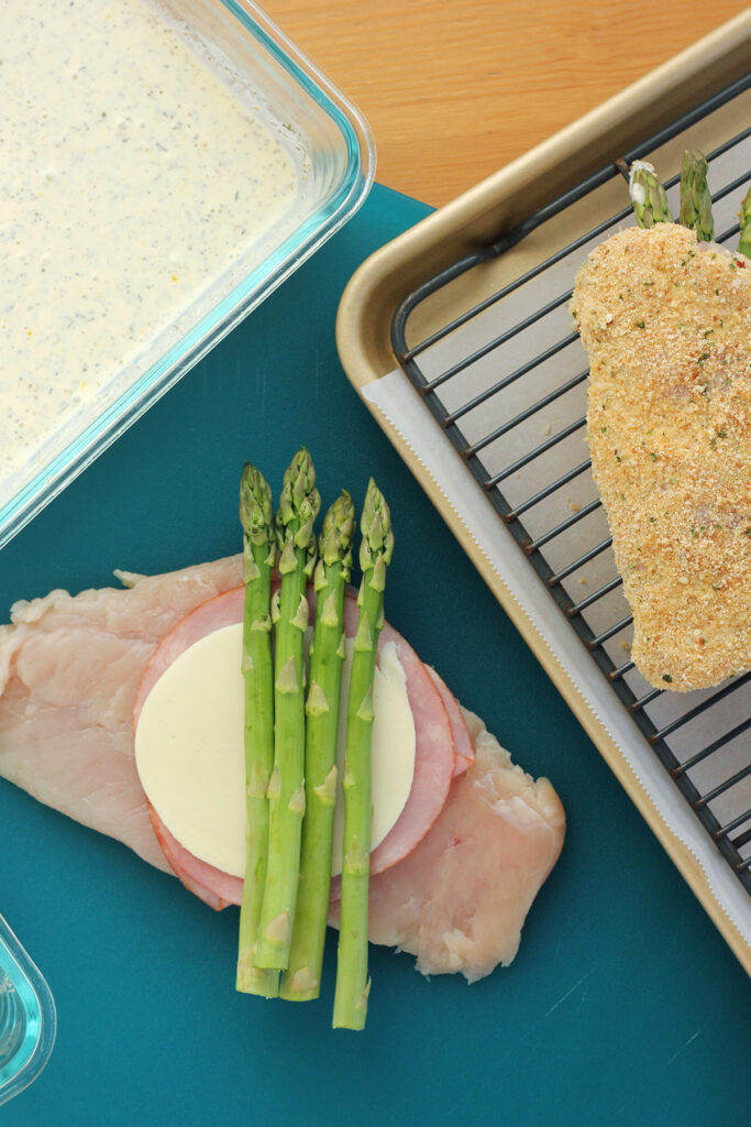blue cutting board with a raw chicken breast that is topped with ham slices, cheese and asparagus.