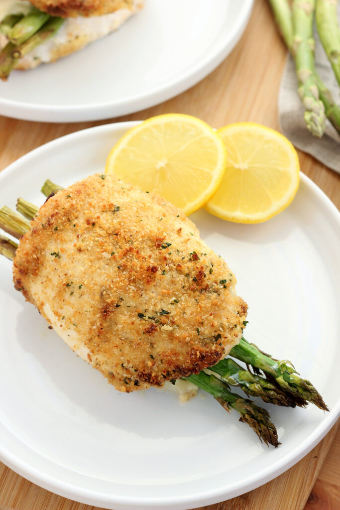 close up image of a golden brown baked stuffed chicken breast with asparagus sitting on a round white plate with two slices of lemon