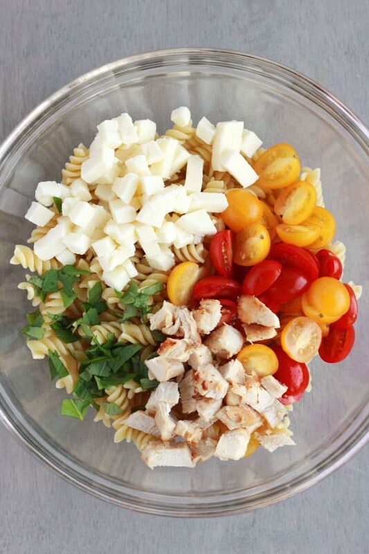 glass bowl filled with cooked spiral pasta and mounds of fresh tomatoes, cooked chicken, mozzarella pieces, and basil.