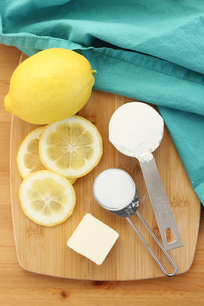 measuring cup filled with flour, spoon filled with sugar, pat of butter and lemon slices sitting on a wooden cutting board with a teal napkin