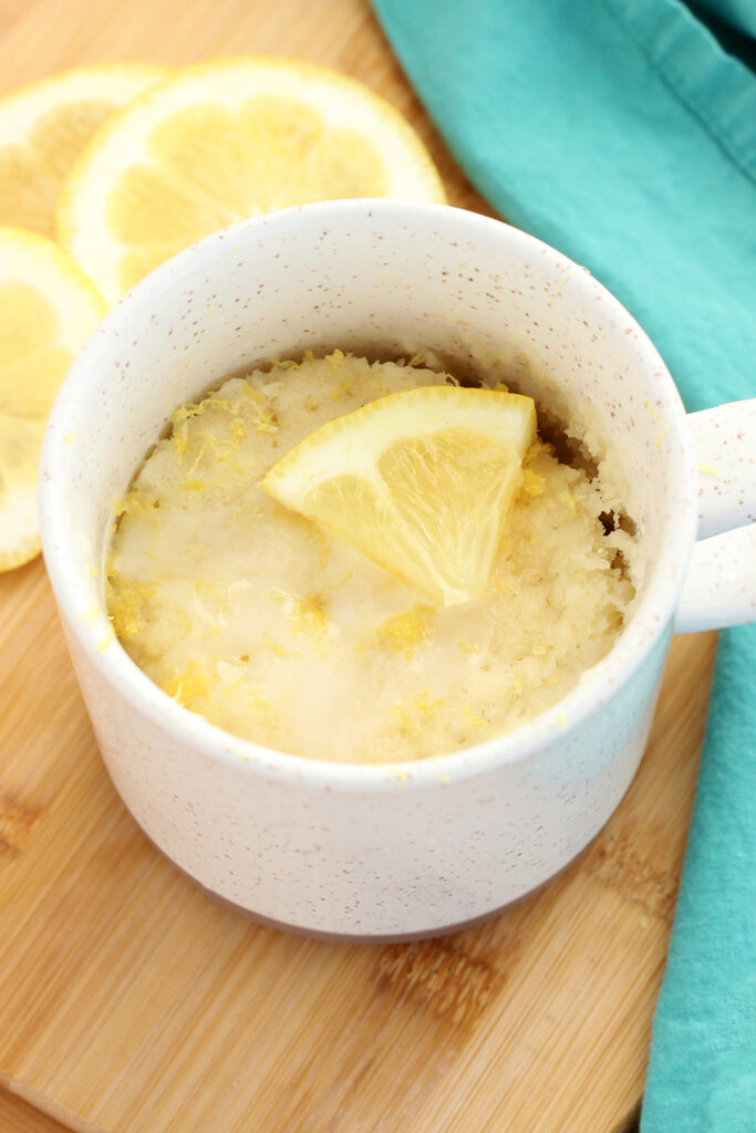 close up image of a tan speckled mug with a cooked lemon cake inside topped with a glaze and fresh lemon wedge