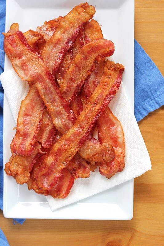 white rectangle plate stacked with a pile of cooked bacon sitting on a blue napkin on a wooden table