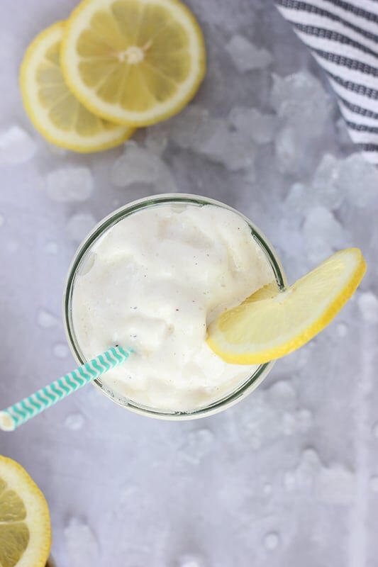 top down image of a mason jar filled with a frozen lemonade with a lemon slice and teal striped straw. Sitting on a grey background with ice pieces and lemon slices
