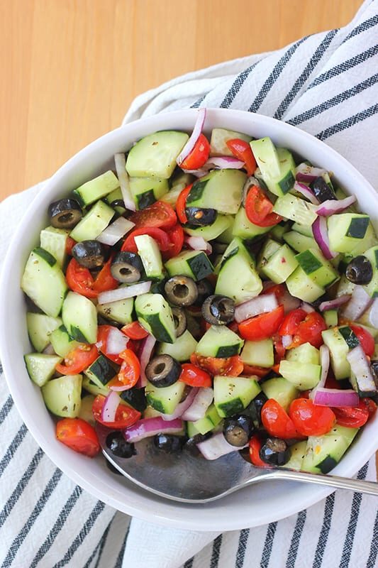 top down image of a white bowl filled with chopped cucumber, tomatoes, red onion, and olives with a spoon off to the side.