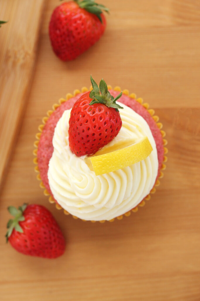 top down image of a pink cupcake that is swirled with white frosting and pressed on a lemon wedge and full strawberry