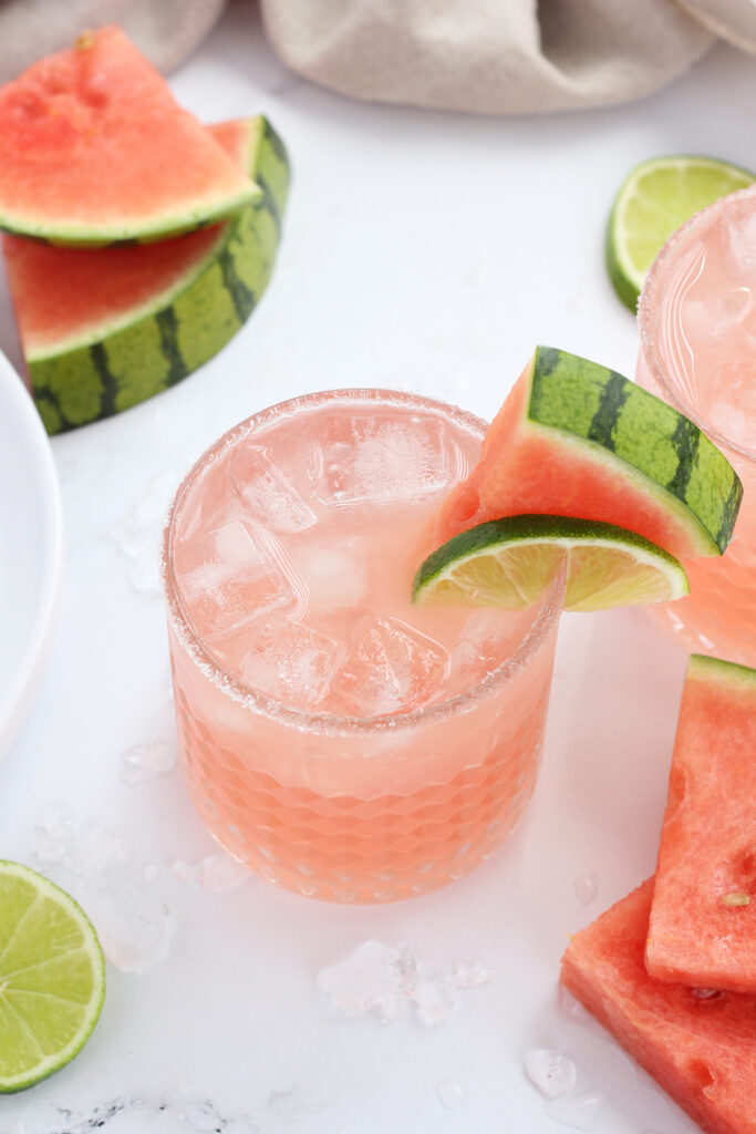 light pink drink in a  sugar rimmed glass with a watermelon slice and lemon slice on the edge