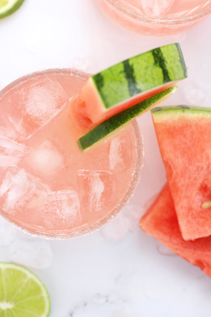 top down image of a pink cocktail with a watermelon wedge on a white table top