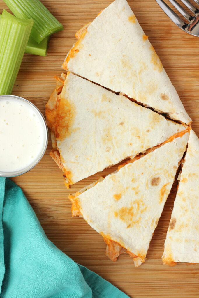 close up of a quesadilla cut into 4 triangles on a wooden cutting board with celery slices and ranch dressing