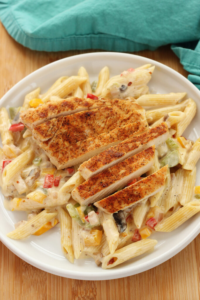 white round plate with a lifted edge filled with pasta that is tossed in a white sauce with brightly colored chopped veggies and a layer of cooked chicken 