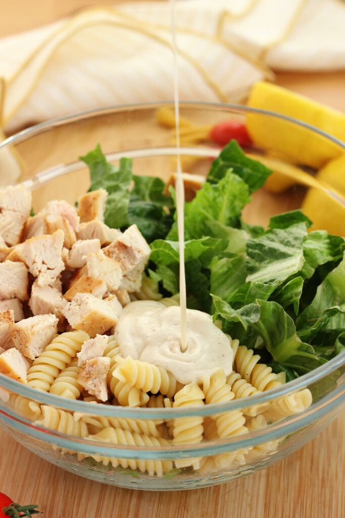 glass bowl filled with lettuce, chicken, and cooked pasta being drizzled with salad dressing