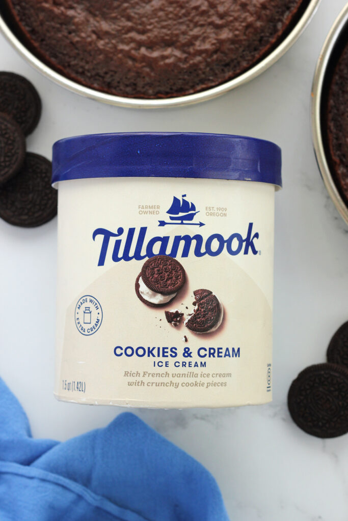 container of Tillamook cookies and cream ice cream on a white table with chocolate cakes off to the side and oreo cookies laid out on the table top