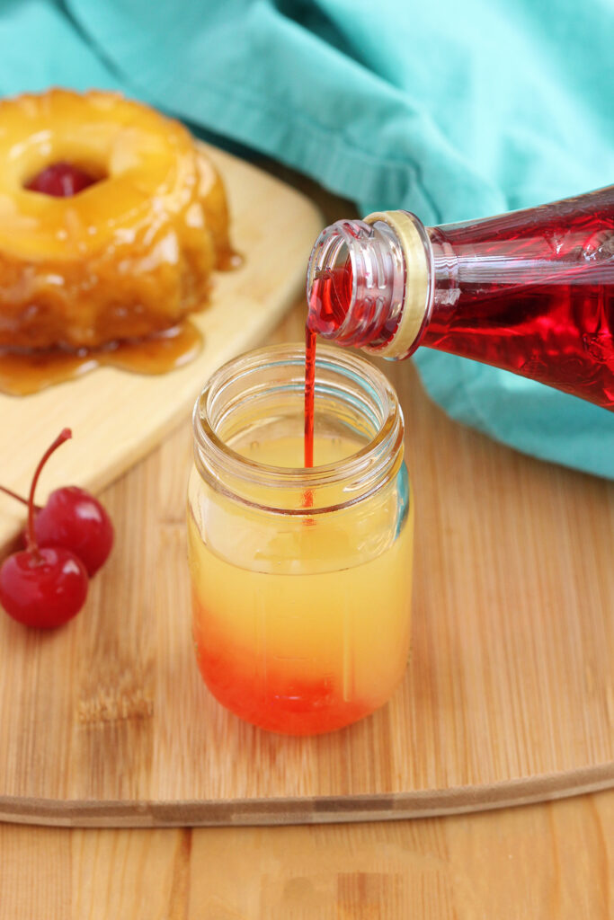grenadine being poured over the top of pineapple juice in a shot glass on a wooden surface