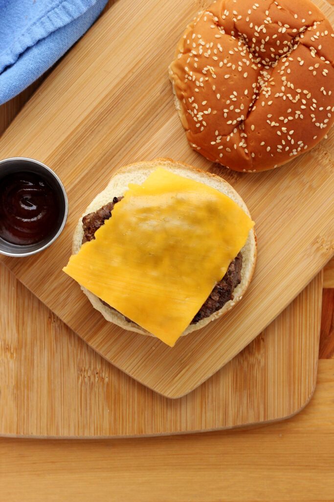 top down image of a burger topped with cheese sitting on a cutting board with a side of bbq sauce in a small metal container