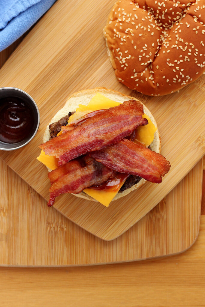 top down image of a burger sitting on the bottom bun with a layer of cheese, bbq sauce and bacon. The top bun is off to the side