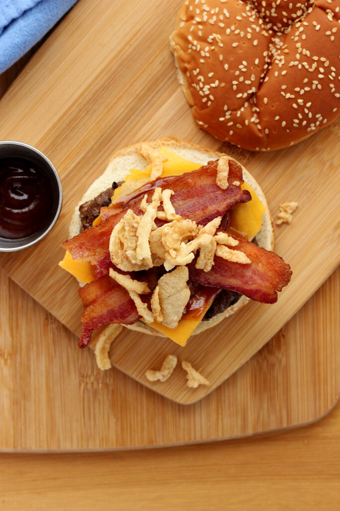 top down image of a burger sitting on the bottom bun with a layer of cheese, bbq sauce and bacon and crispy fried onions. The top bun is off to the side