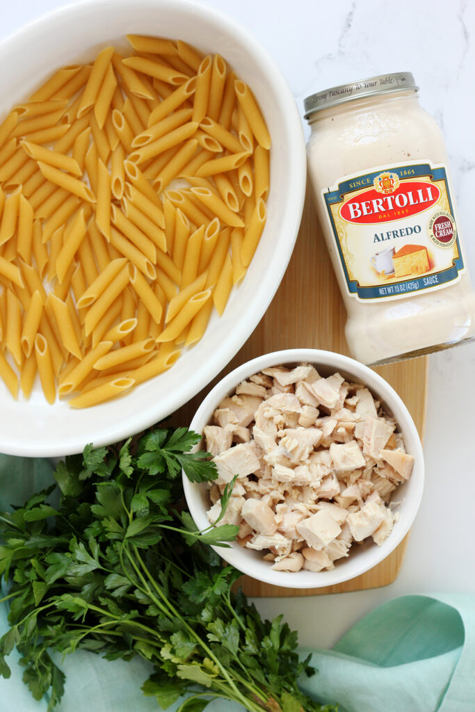 penne pasta in a baking dish, jar of alfredo, diced chicken, and parsley sitting on a wooden cutting board