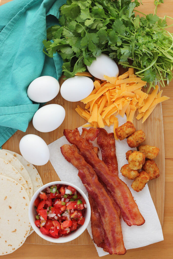 tortillas, bowl of pico, bacon, tater tots, eggs, cheese, and cilantro sitting on a wooden tabletop with a teal napkin