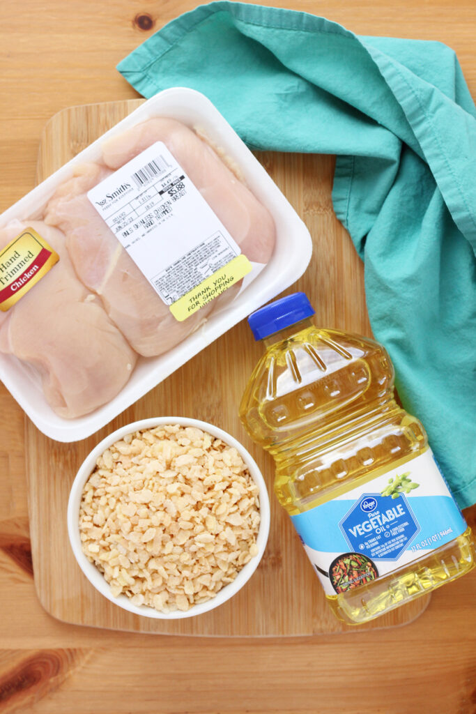 wooden cutting board sitting on a wooden table with a teal napkin off to the side with a package of chicken breast, bowl of rice krispie cereal, and container of vegetable oil sitting on top
