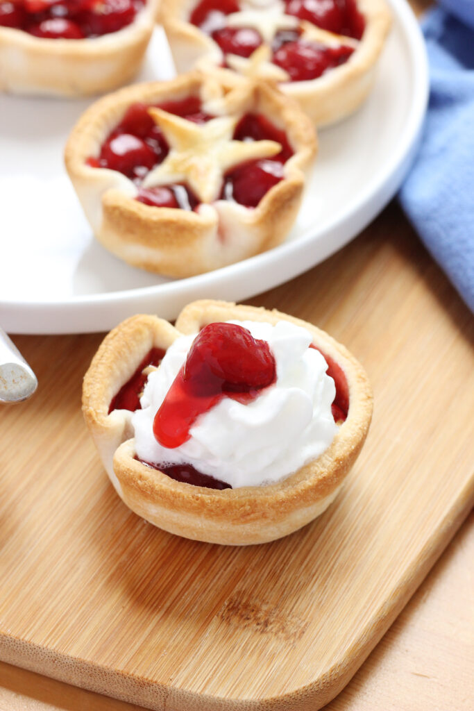 single mini cherry pie topped with whipped cream and a cherry on a wooden cutting board
