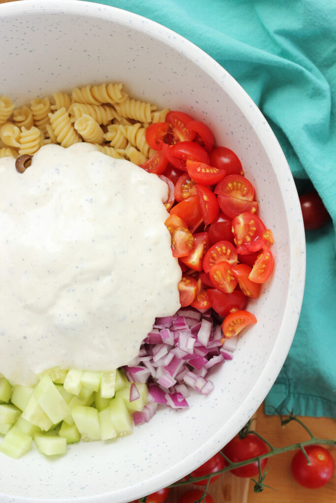 dressing on top of a medley of vegetables and pasta inside a white mixing bowl. 