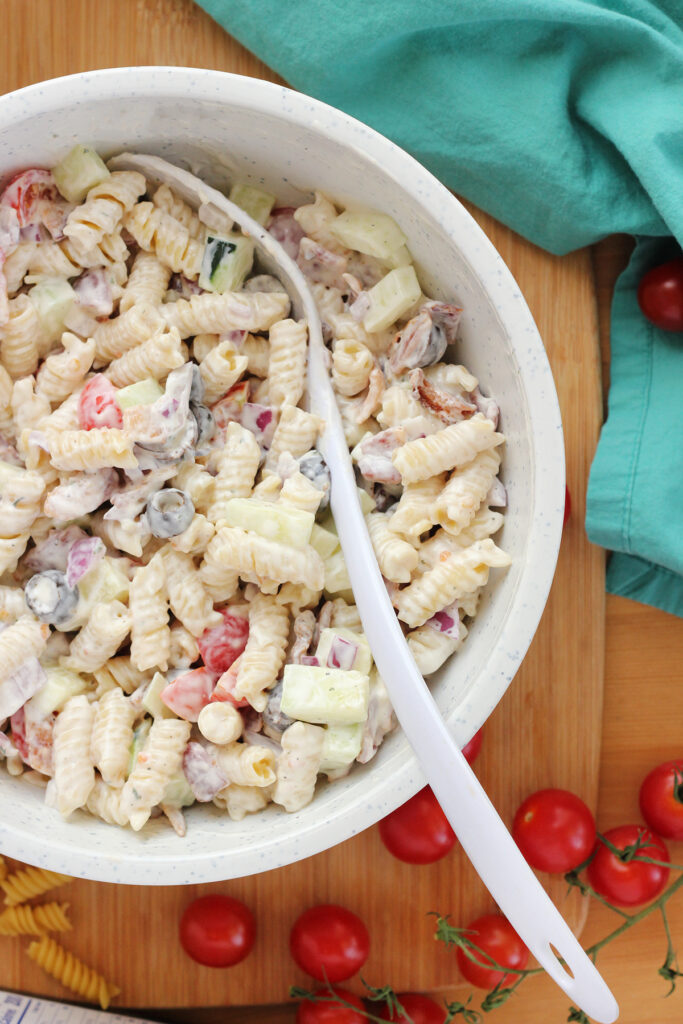 close up of a bowl of pasta salad with a white serving spoon inside the middle. Sitting on a wooden cutting board with cherry tomatoes, dried pasta, and a teal napkin off to the side. 