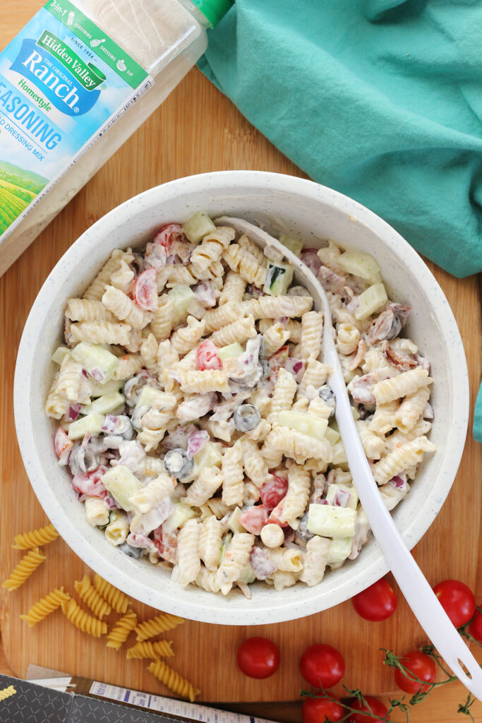 top down image showing a white mixing bowl filled with pasta salad on a wooden cutting board. Around the side are dried pasta pieces, tomatoes, ranch seasoning and a teal napkin