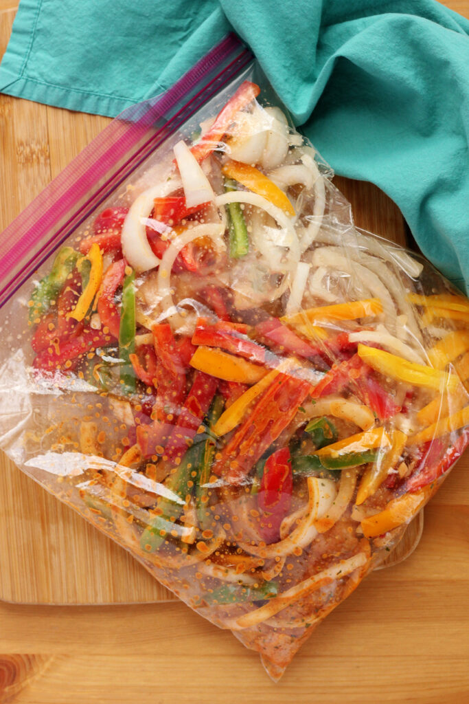 sliced peppers, onion, and chicken inside a large zip top bag with oil and seasonings sitting on a wooden cutting board with a teal napkin off to the side. 