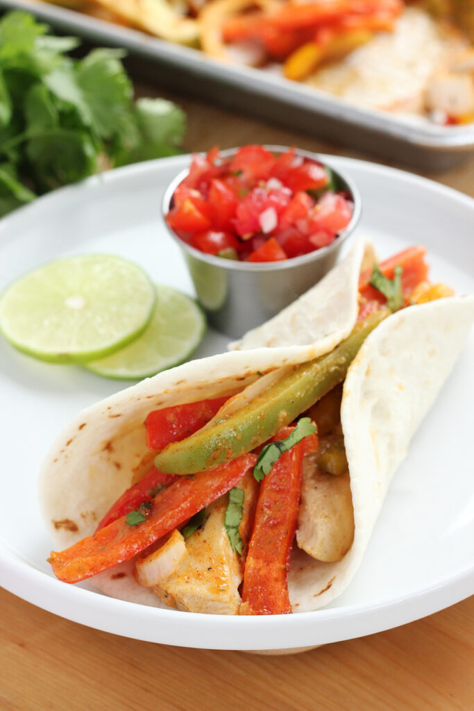 chicken fajitas in a tortilla sitting on a white round plate with a side of chopped tomatoes and lime slices