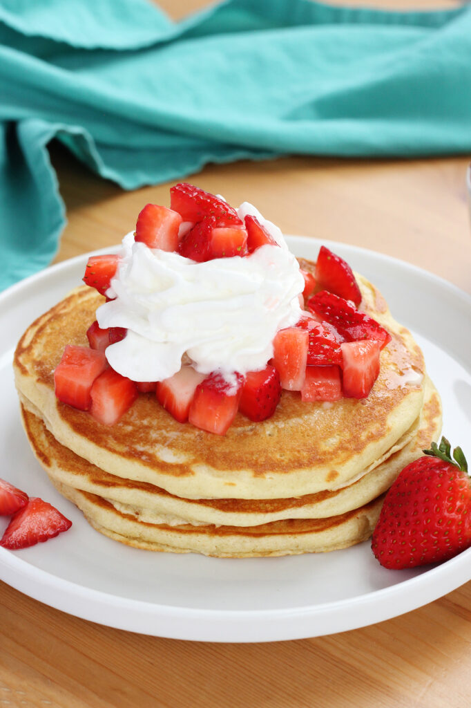 stack of three pancakes sitting on a white round plate topped with chopped strawberries and whipped cream on a wooden cutting board