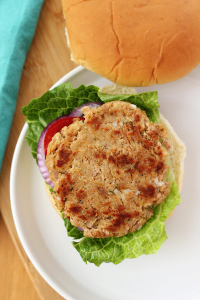 top down image of a tuna patty that has been cooked sitting on the bottom piece of a bun with lettuce, tomato, and onion under