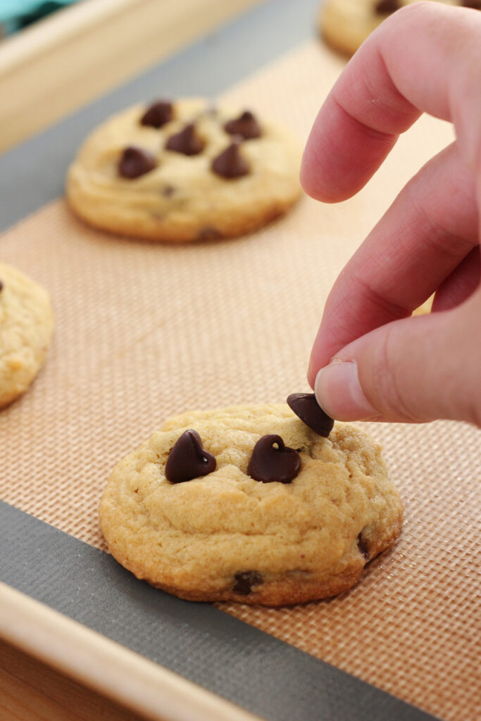 hand placing a chocolate chip onto the top of a cookie that is sitting on a baking tray