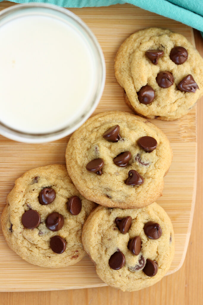 wooden cutting board with a stack of four cookies that have melty chocolate chips on top with a glass of milk on the site
