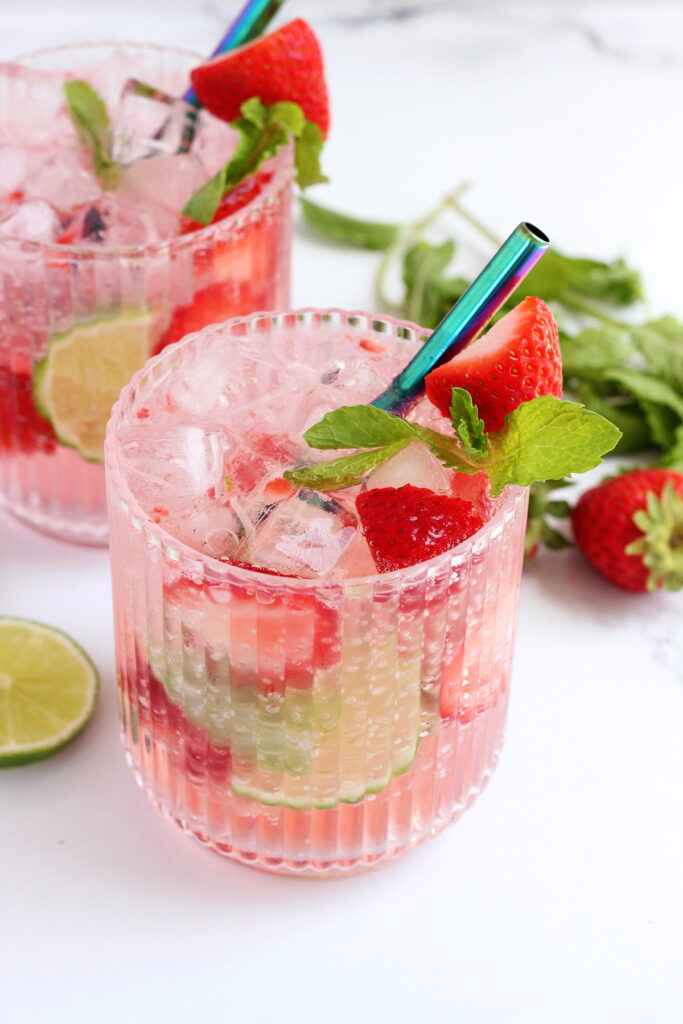 photo of two glasses with ridges filled with pink cocktails that are topped with lime and strawberry slices with mint leaves ona white table