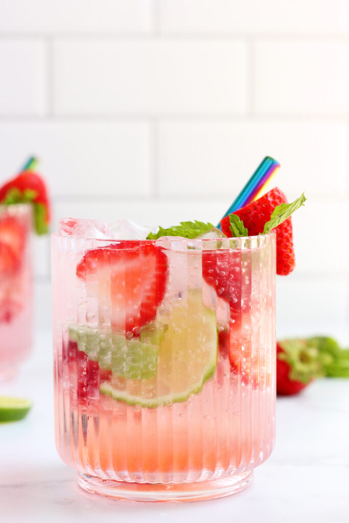 clear glass with ridges filled with a pink cocktail and topped with strawberry slices, lime slices, and pine leaves on a white background