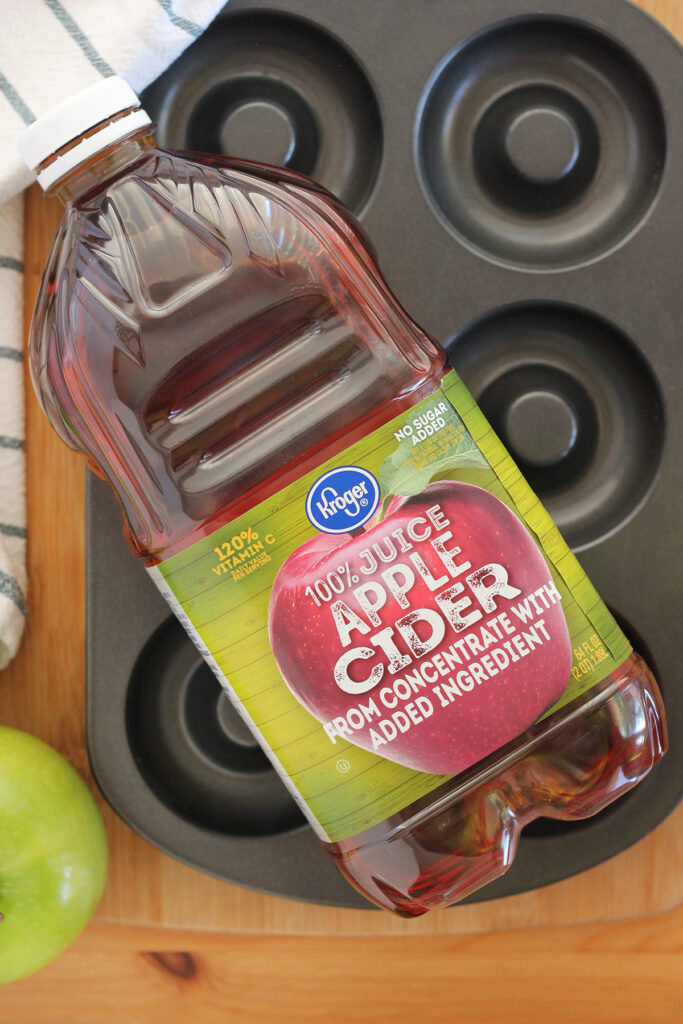 top down image showing a bottle of Kroger apple cider sitting on top of a donut baking pan on a wooden table top with a green apple off to the side