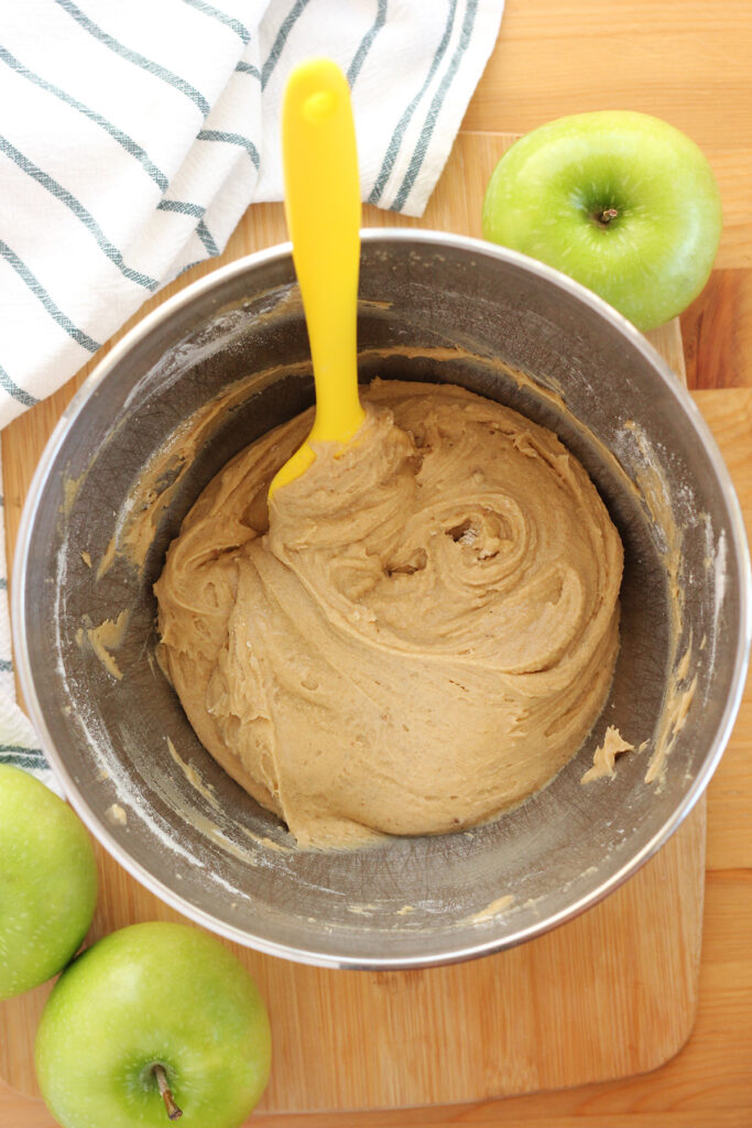 top down image of a brown cake batter in a kitchen aid mixing bowl with a yellow spatula sitting inside. The bowl is sitting on a wooden table top with a stripe napkin and green apples off to the side