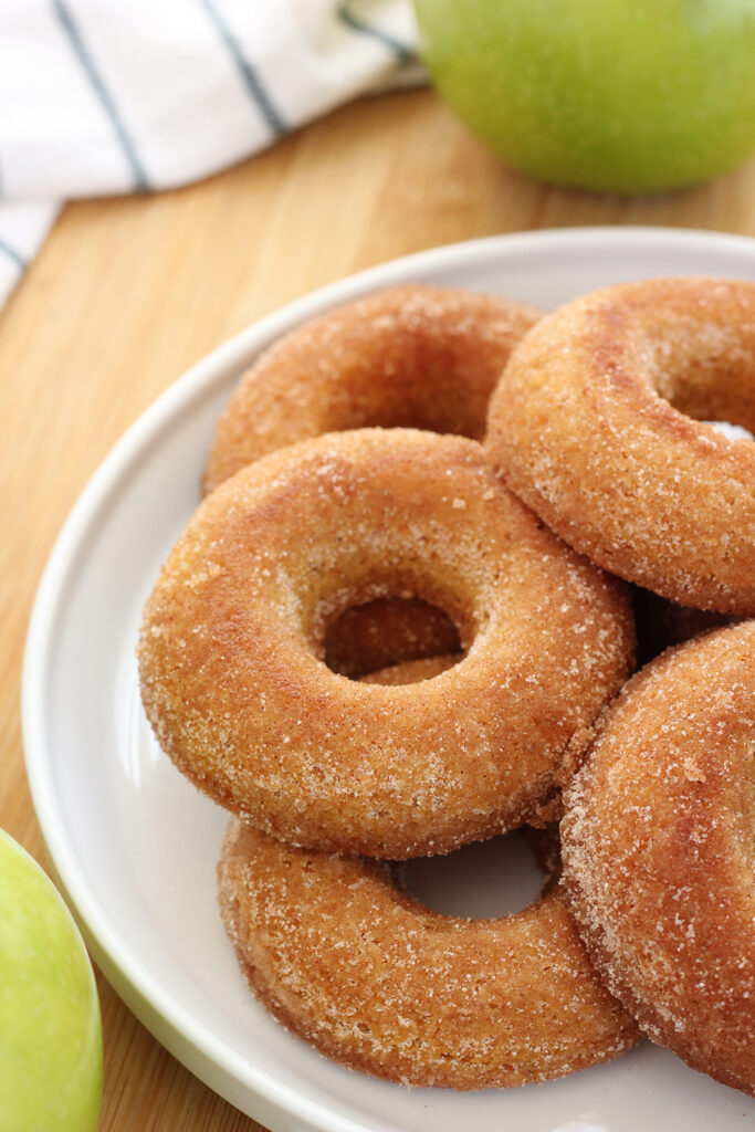 small round white plate with a lifted edge topped with a pile of sugar coated donuts. green apples and a white and blue stripe napkin off to the side