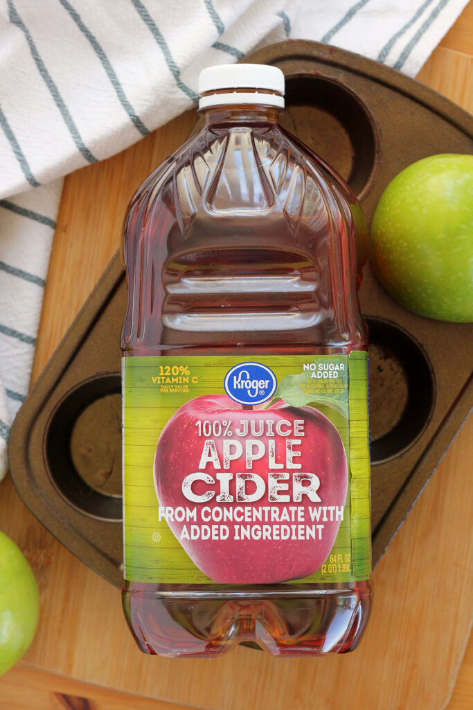 top down image of a bottle of apple cider sitting on a six count muffin tin with green apples off to the side on a wooden table top