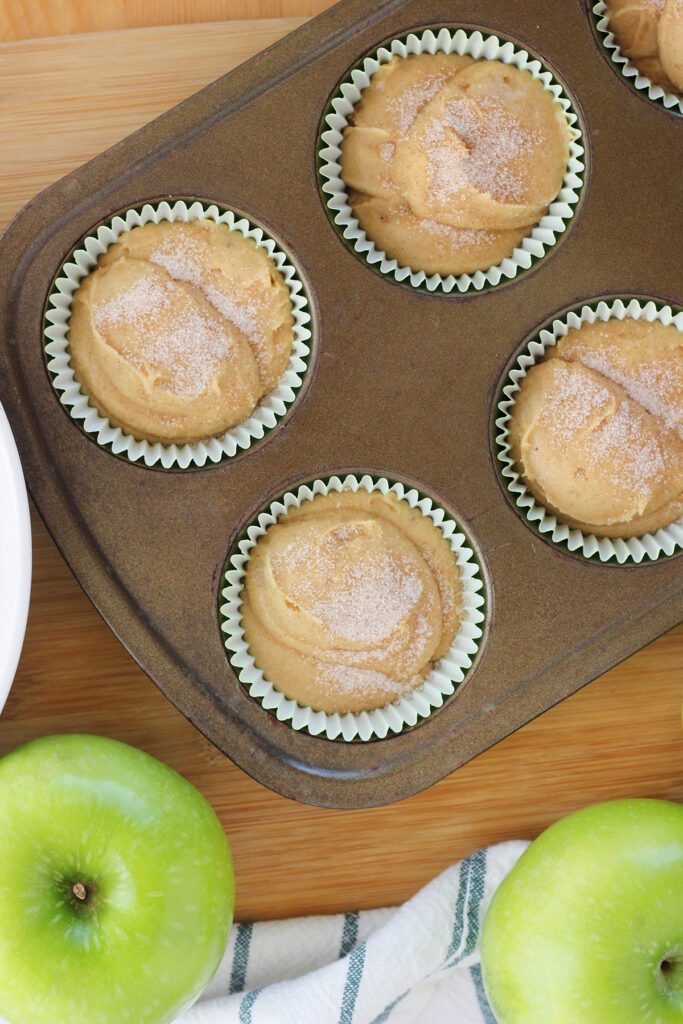 top down image of a six count muffin tin wtih unbaked muffins that have been sprinkled with cinnamon and sugar. the pan is on a wooden table top with green apples and a white and green striped napkin off to the side